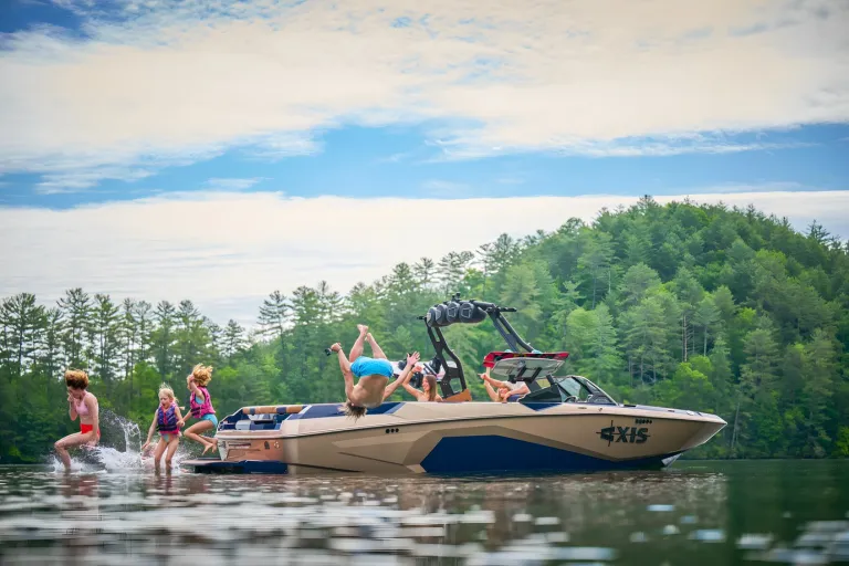 Friends leap and backflip into the water off a navy-and-tan Axis A245 near a forested shoreline under a blue sky.