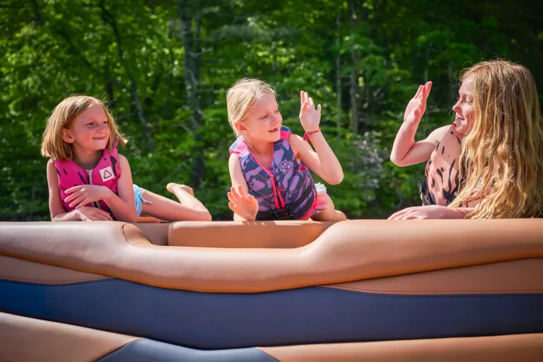 Three young girls in life vests laugh and high-five in the tan bow seating of an Axis A245, forest behind them.