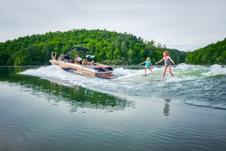 A navy-and-tan Axis A245 tows a wake foiler across a glassy, tree-lined lake, passengers watching from the cockpit.