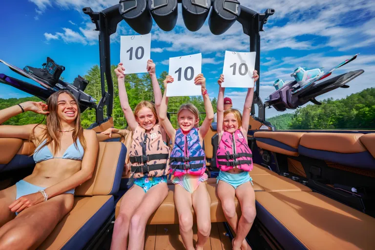 A woman and three girls in life vests hold up '10' judging scorecards from the tan interior of an Axis A245 on a sunny lake.