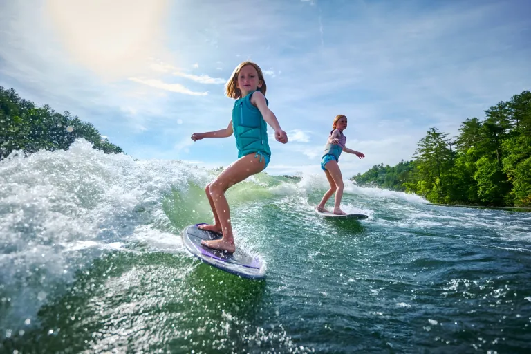 Two young girls wakesurf side by side on a sunlit wave behind an Axis A245, forested shoreline beyond.