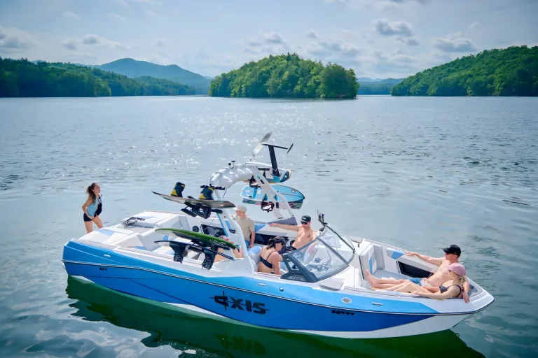 A white-and-blue Axis T250 idles on a calm lake before a forested island, friends aboard and one on the swim platform under a blue sky.