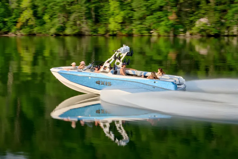 A white-and-blue Axis T250 runs across a calm lake with passengers aboard, green forested shore reflected in the water.