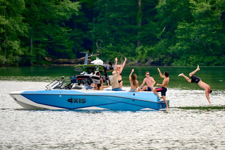Friends cheer aboard a white-and-blue Axis T250 near a forested shore as one person backflips into the water.