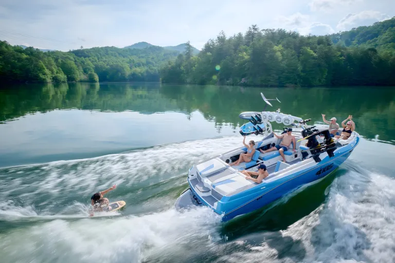A white-and-blue Axis T250 tows a wakesurfer across a calm, misty lake ringed by forested hills.
