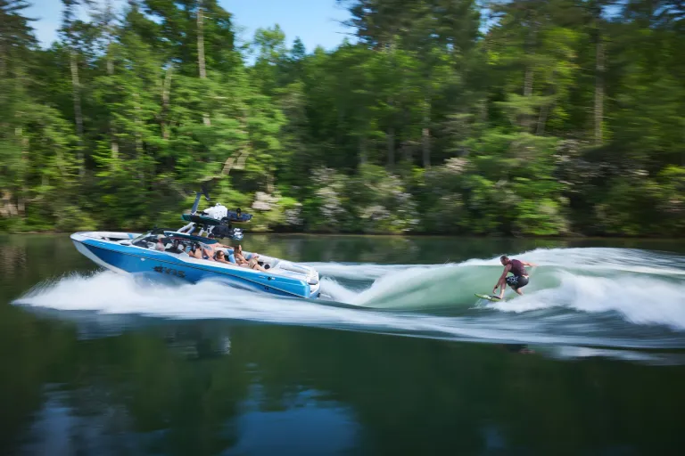 A white-and-blue Axis T250 tows a wake foiler along a forested shoreline, spray trailing behind.