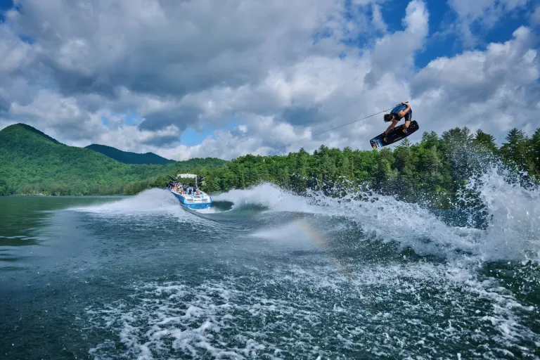 A wakeboarder boosts huge air off the wake behind a white-and-blue Axis T250, a rainbow glinting in the spray under a cloudy sky.
