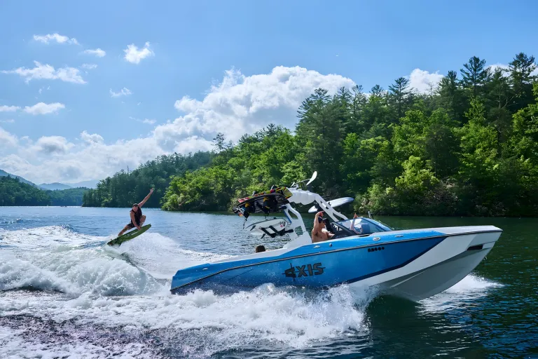 A white-and-blue Axis T250 runs in side profile as a wakesurfer rides its wake, forested shoreline and blue sky behind.