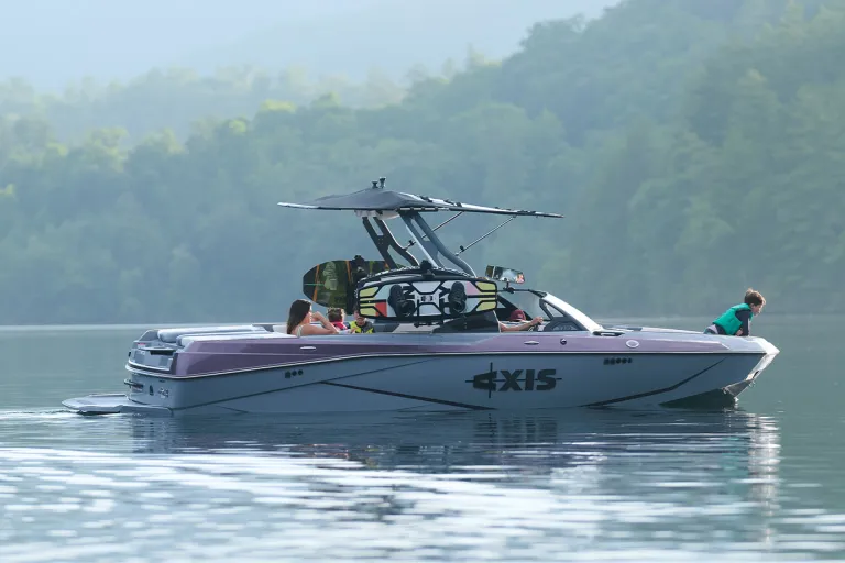 An Axis T220 sits in full side profile on misty, glassy water, family aboard and wakeboards stowed on the tower rack against forested hills.