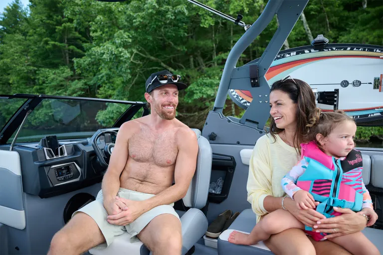 A man and a woman holding a toddler smile while seated in the Axis T220 cockpit beside the helm, a retro-striped wakeboard racked on the tower behind them.