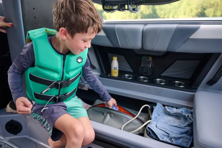 A boy in a teal life vest reaches into the Axis T220's open under-seat storage, packed with a gear bag, clothing and sunscreen.