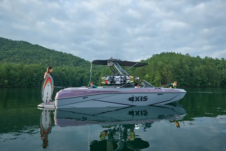 A woman stands on the swim platform holding a wakesurf board beside a docked Axis T220, kids seated aboard with calm water and forested hills behind.