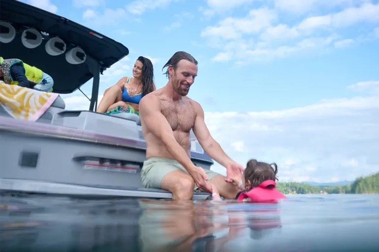 A man sits on the Axis T220 swim platform, lowering a small child in a pink life vest into the water while a woman relaxes aboard on a sunny day.