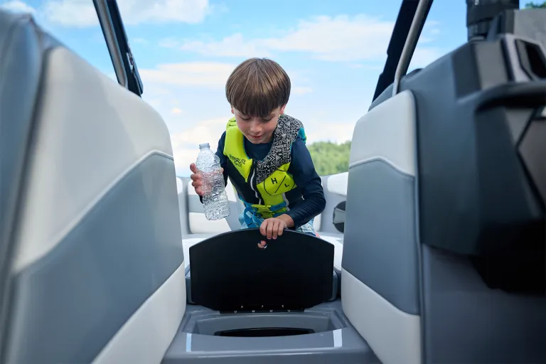 A young boy in a life vest holds a water bottle while lifting the lid of the Axis T220's in-floor trash can under a bright blue sky.