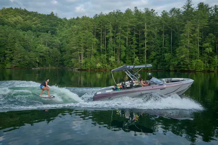 A woman wakesurfs behind an Axis T220 as family members ride aboard, the boat shown in full side profile on a tree-lined lake.