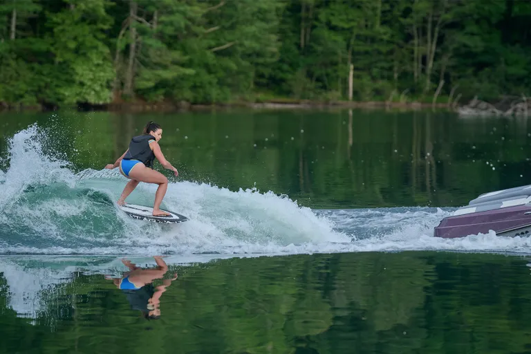 A woman wakesurfs the clean wake behind an Axis T220, carving across glassy green water with forested hills in the background.
