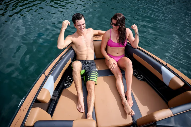 A young couple lounges in the Axis T235's spacious bow seating, tan cushions and cup holders around them and green water alongside.