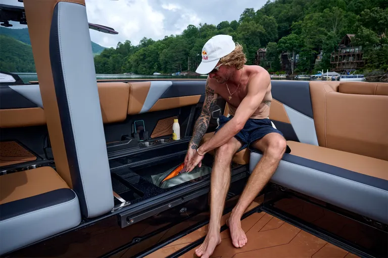 A man reaches into the Axis T235's in-floor cooler storage from the tan rear lounge, lakefront homes and forested hills in the background.