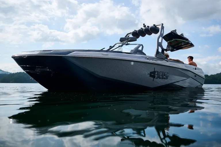 An Axis T235 rests on calm water in a three-quarter bow view, two riders at the stern beneath a cloudy sky with forested mountains behind.