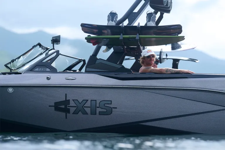 A man relaxes aboard an Axis T235 shown in side profile, wakeboards and a surfboard loaded on the tower rack against hazy mountains.
