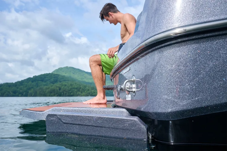 A man sits on the Axis T235's swim platform with his feet on the step, calm water and a green mountain shoreline stretching behind him.
