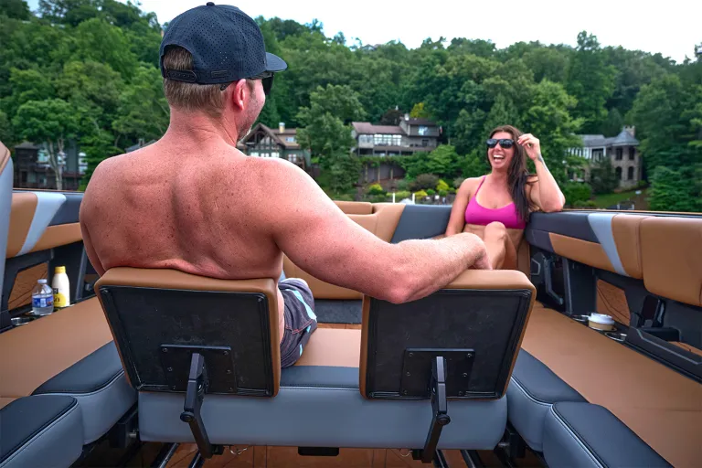 A man sits on the Axis T235's rear-facing WakeView bench talking with a woman across the cockpit, lakefront homes and trees behind them.