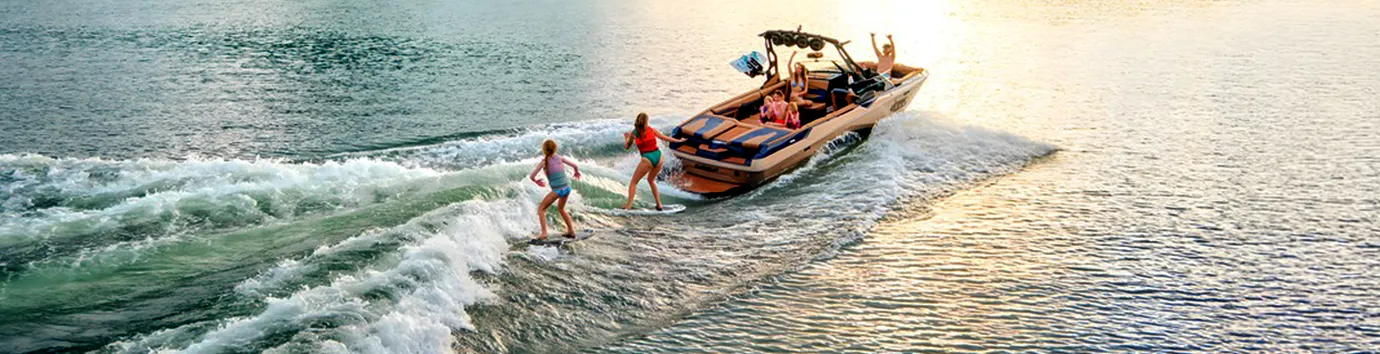 Two young wakesurfers ride the wake behind a bronze Axis A245 as family cheers from aboard, a green mountain and sunset sky behind them on the lake.