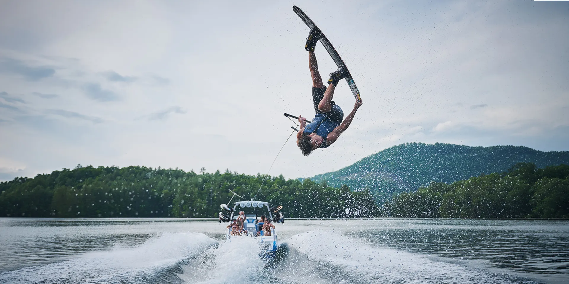 A wakeboarder flips high above the wake behind a fully loaded Axis wake boat, the crew watching on a mountain lake.
