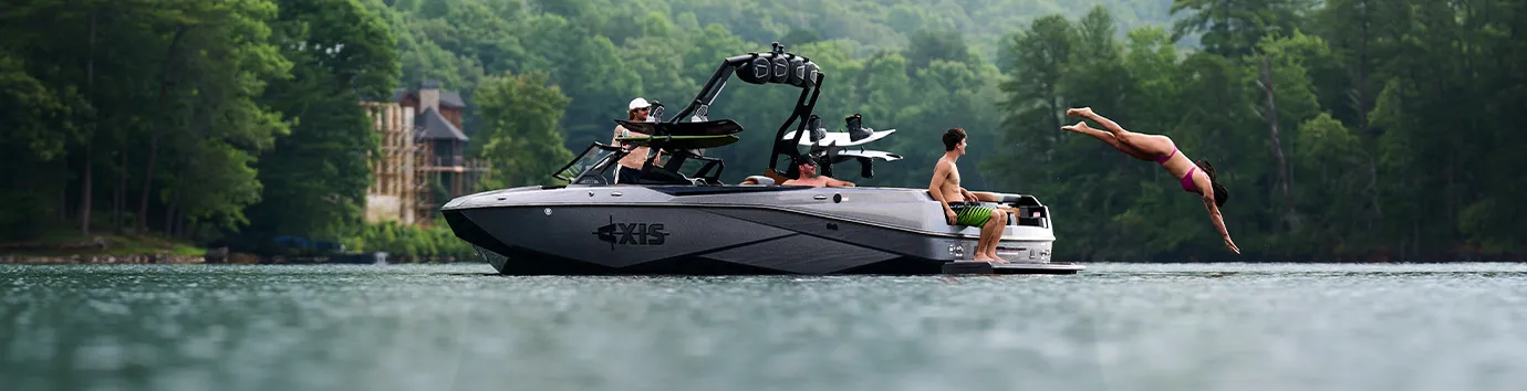 A woman dives off the swim platform of a gray Axis T220 into a calm lake as three people relax aboard, wooded hills and a lakeside home behind.